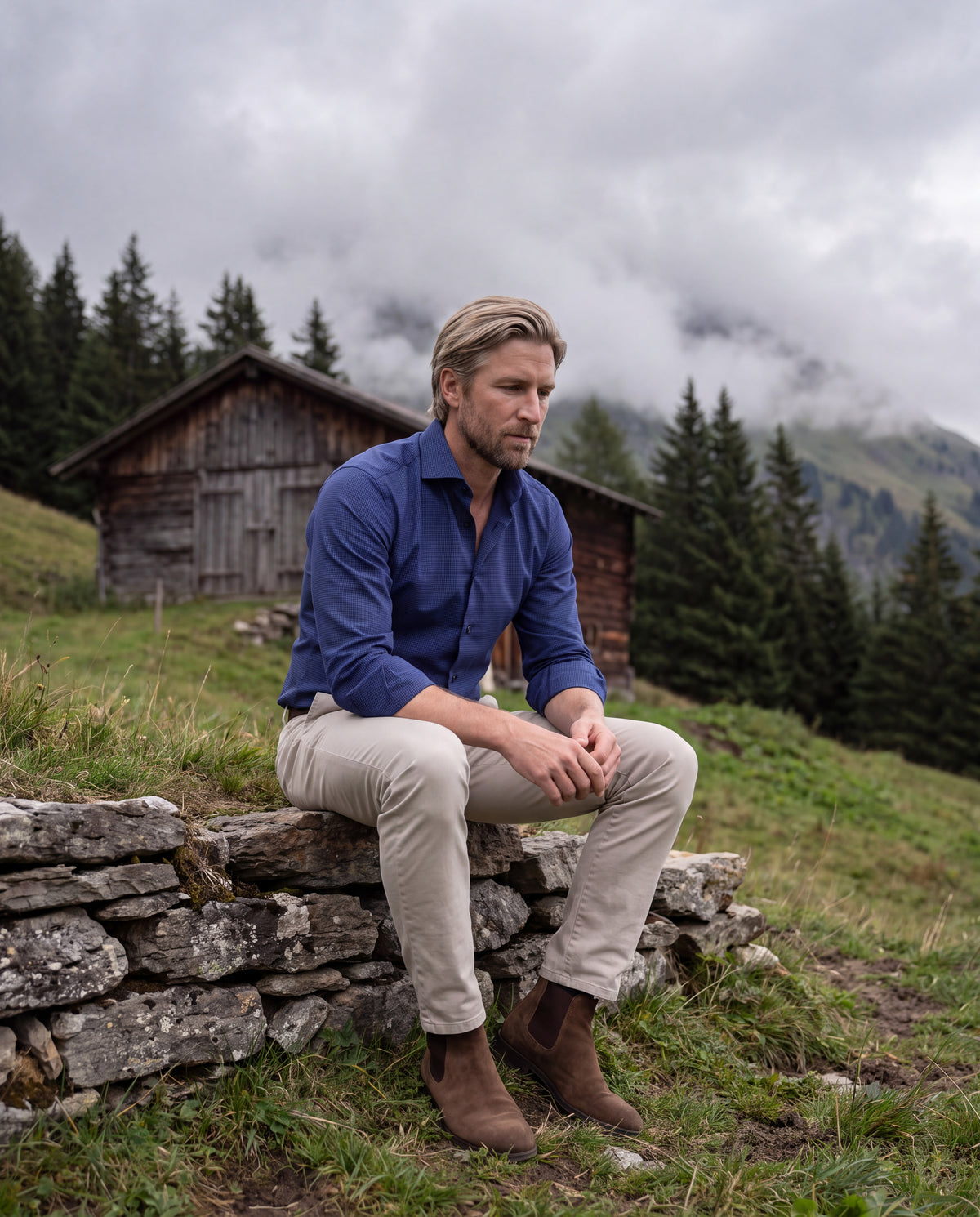 Mann im blauen Einhorn Hemd auf einer Steinmauer vor einer Alphütte